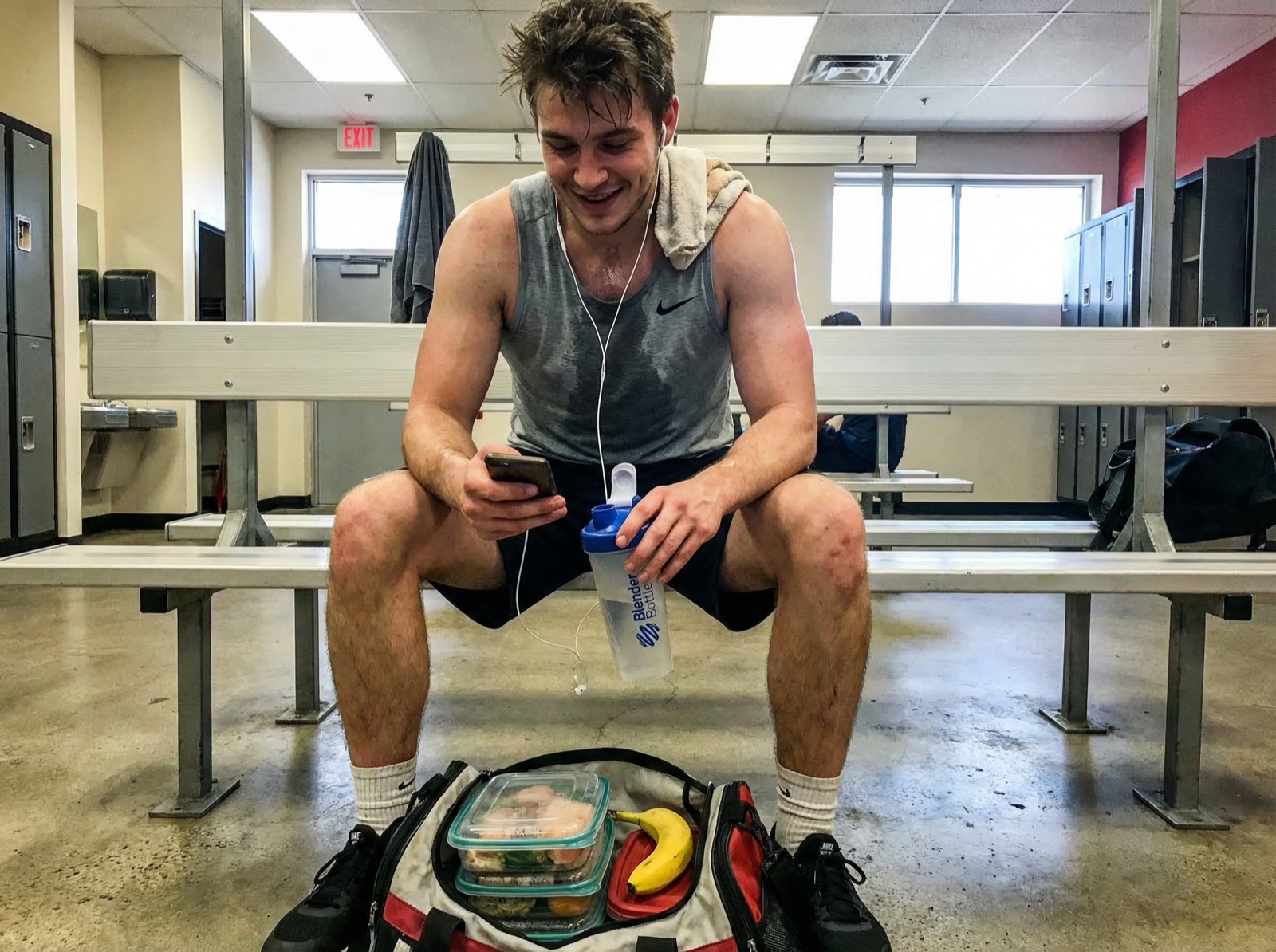 Guy in a Nike tank top sitting on a gym bench post-workout, scrolling his phone with a BlenderBottle between his knees, gym bag with meal prep containers on the floor